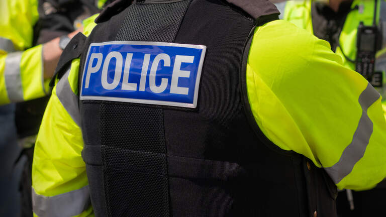 Close-up of 'POLICE' marking written on the back of a hi-visibility stab proof vest worn by a trio of police officers at the scene of an incident. Officers enforce the law, establish public order and protect citizens.