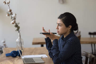 Serious focused Indian woman lead talk on speakerphone sit workplace desk with laptop holds smartphone near mouth calling to client, explain details to customer working at home office use modern tech